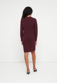 Donna con lunghi capelli scuri che sta di spalle, indossa un vestito aderente a maniche lunghe di lunghezza ginocchio di colore borgogna e sandali neri con tacco.