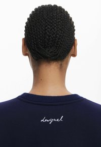 Marinblaues Sweatshirt mit geripptem Kragen, das ein weißes gesticktes "desigual"-Logo auf dem Rücken trägt. Texturiertes, geflochtenes Haar ist sichtbar.