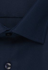 Marineblauwe polo in een gestructureerde stof, met een klassieke kraag en een zwarte placket met vier knopen. Close-up van de kraag en de knoopdetails.