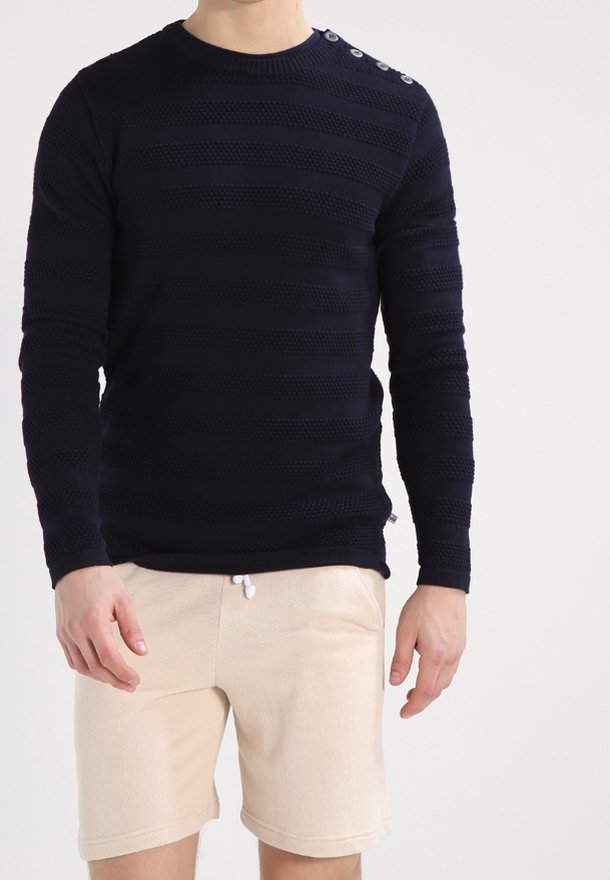 Homme portant un pull à manches longues bleu marine texturé avec des détails de boutons sur l'épaule et un short beige à cordon de serrage.