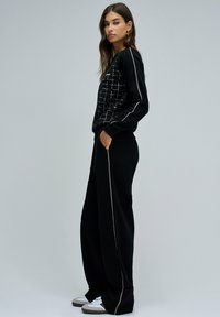 Sweatshirt noir avec un devant texturé et à motifs, associé à un pantalon noir ample avec un détail de fermeture éclair sur le côté. Baskets blanches visibles.