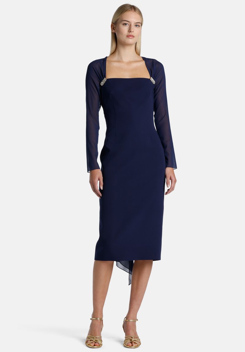 Tamsiai mėlyna suknelė su struktūruotu kvadratiniu iškirpimu, ilgomis permatomomis rankovėmis ir dekoratyviniais sidabriniais akcentais ant petnešėlių. Vidutinio ilgio dizainas.