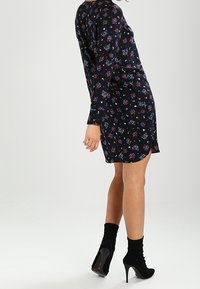 Květinové šaty s dlouhými rukávy, navy modré pozadí, červené a bílé květy, délka nad kolena, doplněné černými kotníkovými botami a podpatky.