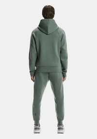 Zöld kapucnis pulóver nagy kapucnival és bordázott szegéllyel, hozzáillő tapered (keskenyedő) melegítőnadrággal párosítva. Az anyag puhanak és sima textúrájúak tűnik.