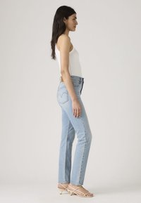 Lichtblauwe recht gesneden jeans met hoge taille, met subtiele achterste stitching en een bruine leren patch, gecombineerd met beige sandalen met bandjes.