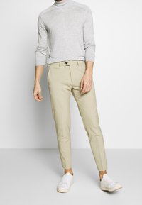Jasnoszary sweter z golfem, beżowe spodnie garniturowe z zapinaniem na guziki i białe sneakersy. Smukły design, dopasowany krój, minimalna tekstura.