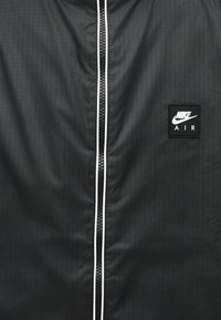 Μαύρο μπουφάν Nike Air μπροστά με κεντρικό μαύρο φερμουάρ περιγραμματισμένο με λευκό και σήμα λογότυπου Nike Air στη δεξιά πλευρά.