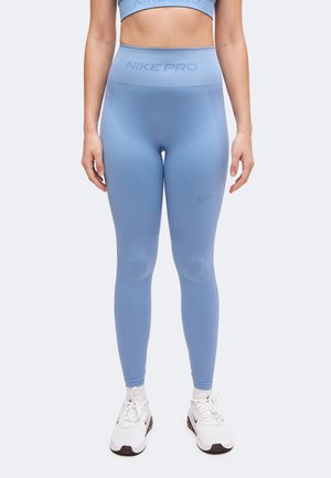 Kvinde iført lyseblå Nike Pro leggings med høj talje og matchende sports-bh med hvide Nike sneakers, stående mod en neutral baggrund.