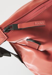 Sac imperméable rouge avec fermeture éclair, doté d'une sangle et d'un détail texturé. Le nom de la marque "RAINS" est embossé sur le tirette de la fermeture éclair.