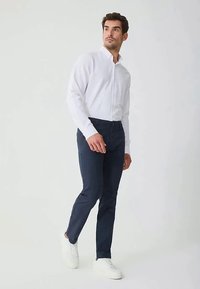 Vīrietis iet, ģērbies baltā krekla ar pogām, tumši zilos biksēs un baltos sporta apavos, uz vienkārša gaiša fona.