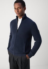 Cardigan zippato a coste blu navy con colletto alto, tasche laterali, tessuto testurizzato e un logo nero sulla manica sinistra.
