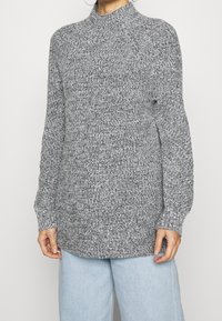 Grå stickad tröja med hög krage, långa ärmar och ribbad mudd. Texturen innehåller en blandning av ljusa och mörka grå garn.