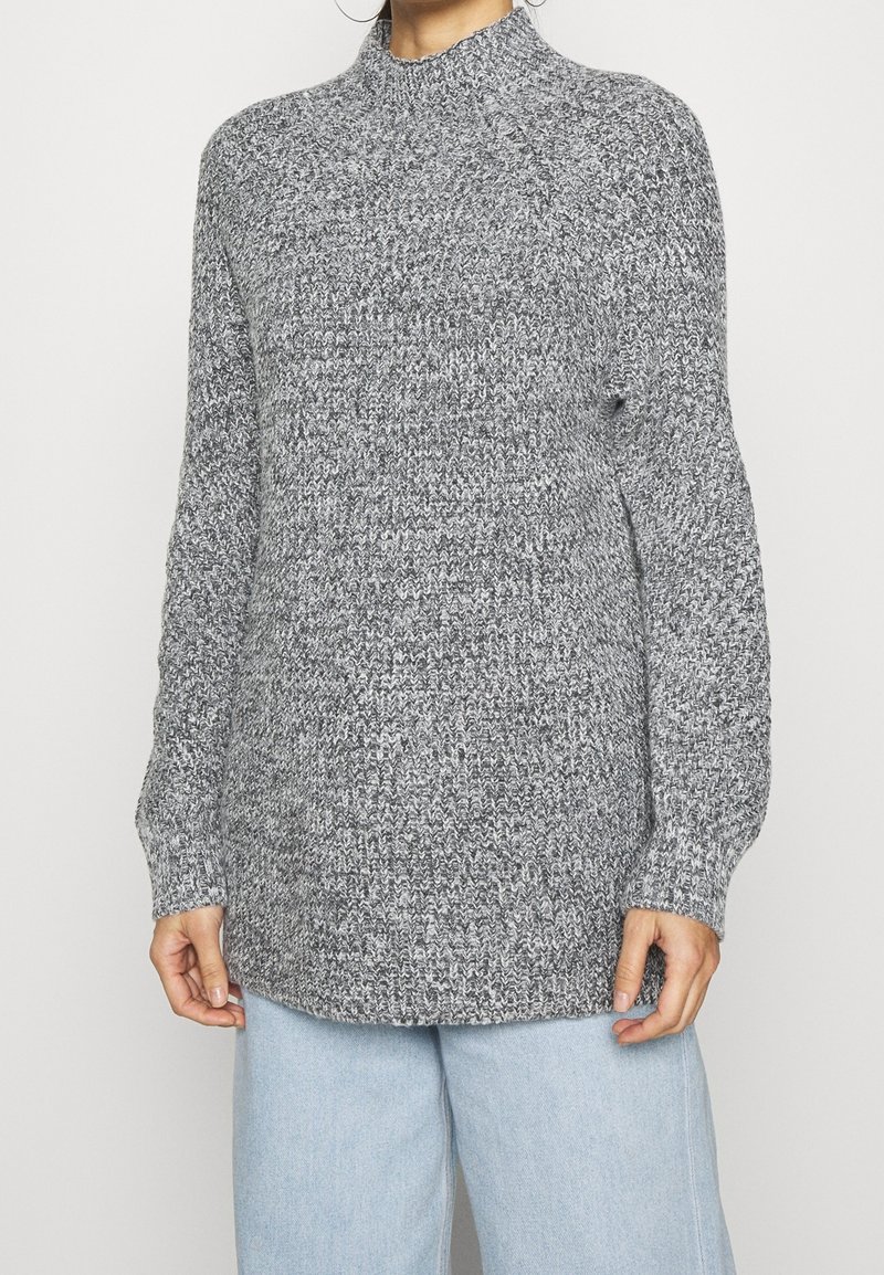 Grå stickad tröja med hög krage, långa ärmar och ribbad mudd. Texturen innehåller en blandning av ljusa och mörka grå garn.