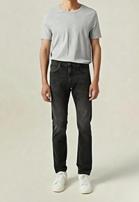 Personne portant un t-shirt gris clair à manches courtes, un jean noir et des baskets blanches, debout devant un fond neutre.
