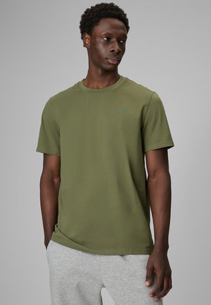 Mężczyzna ubrany w gładki oliwkowo-zielony t-shirt z krótkim rękawem z małym logo na piersi i jasnoszare spodnie dresowe na jednolitym tle.