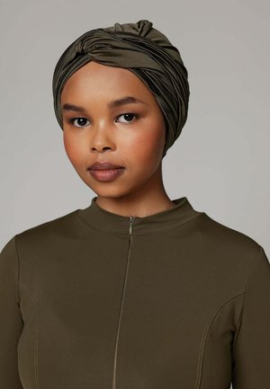 Jonge vrouw met een gladde bruine huid draagt een olijfgroene hoofdband en een bijpassende top met rits, neutrale achtergrond.