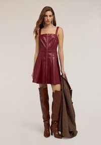Donna che indossa un vestito in pelle bordeaux senza maniche con zip frontale e stivali marroni fino al ginocchio, mentre tiene un cappotto a scacchi in mano.