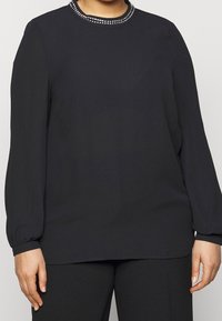 Svart långärmad blus med rund halsringning som har en dekorativ rad av små silvernitar längs kragen. Slät tygtextur.