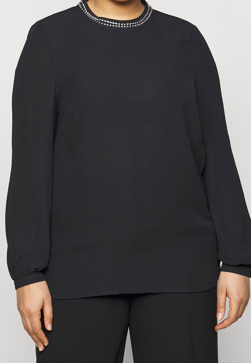 Svart långärmad blus med rund halsringning som har en dekorativ rad av små silvernitar längs kragen. Slät tygtextur.