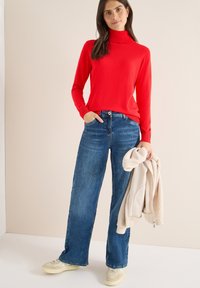 Pull à col roulé rouge assorti à un jean bleu à jambes larges. Tenant une veste polaire beige. Porte des baskets de couleur claire. Textures lisses affichées.