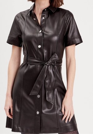 Femme portant une robe boutonnée en cuir noir à manches courtes avec une ceinture nouée à la taille, debout devant un fond clair uni.