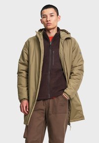 Jeune homme portant une veste polaire marron sous un manteau d'hiver à capuche vert olive ouvert avec un pantalon marron, debout avec une main dans la poche.
