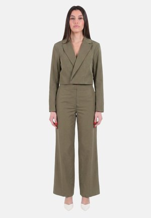 Donna in piedi che indossa un blazer corto verde oliva e pantaloni a gamba larga e vita alta abbinati con scarpe bianche appuntite.