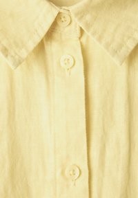 Camicia di lino gialla con colletto classico, sei piccoli bottoni e un tessuto testurizzato, dall'aspetto sottile e leggero.