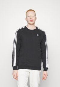 Sweatshirt noir avec un design à trois rayures blanches, poignets et ourlet côtelés, arbore un petit logo blanc sur la poitrine. Texture lisse, coupe décontractée.