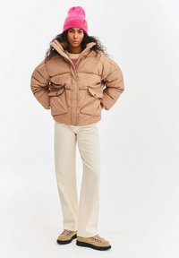 Béžová prešívaná bunda s textúrou a štyrmi prednými vreckami, v kombinácii s krémovými širokými nohavicami a béžovými turistickými topánkami. Jasne ružová čiapka.