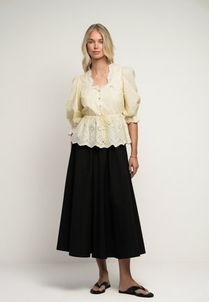 Blondine vrouw met een gele blouse met pofmouwen van oogjeskant en een zwarte midirok, staand tegen een effen lichte achtergrond.