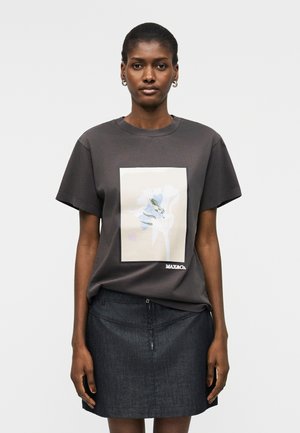 Naine, kes kannab tumedat halli t-särki, millel on valge lilleline kujundus ja tekst "MAX&Co.", koos tumeda teksaseelikuga.