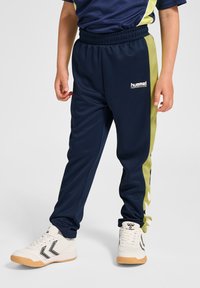 Granatowe spodnie dresowe z zielonymi paskami po bokach, elastycznym pasem i logo. Dziecko nosi białe sneakersy z czarnymi i beżowymi detalami na podeszwie.