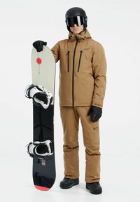 Cazadora y pantalones de snowboard marrones, guantes negros, sosteniendo una tabla de snowboard negra con fijaciones blancas y acentos rojos, gafas de nieve en el casco.