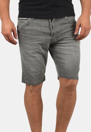 Homme portant un short en jean gris délavé avec braguette à boutons et poches latérales, debout devant un fond blanc uni.