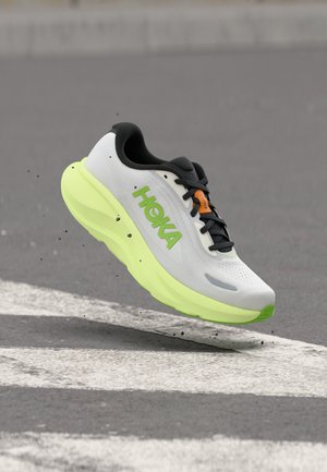 Zapatilla de running blanca y verde neón con cordones negros y logo de HOKA, suspendida sobre asfalto con una línea blanca pintada.