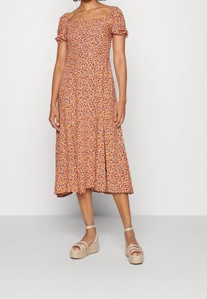 Femme portant une robe midi orange rouille à petits motifs floraux blancs et jaunes, avec des manches bouffantes, associée à des sandales compensées beige.