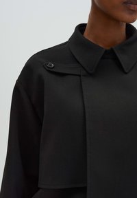 Zwarte jas met een gestructureerd ontwerp, voorzien van een puntkraag, enkelvoudige knoopsluiting en een gladde, matte textuur.