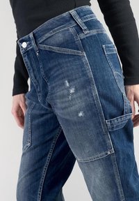 Persona que lleva jeans de mezclilla de retales azul oscuro con detalles desgastados y una camiseta de manga larga negra con textura en relieve, de pie contra un fondo liso.