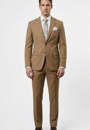 Hombre de pie con un traje color beige, camisa blanca, corbata de cachemira, pañuelo blanco en el bolsillo y zapatos de vestir marrones, frente a un fondo liso.