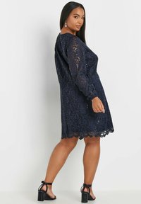 Vestido de encaje azul marino con mangas largas y dobladillo con forma de concha. La superposición de encaje transparente revela una silueta ajustada debajo. Sandalias de tacón completan el look.