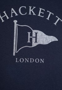 Mørkeblå bomull T-skjorte med et hvitt silkscreen trykk av et flagg med en "H" og teksten "HACKETT LONDON" under.