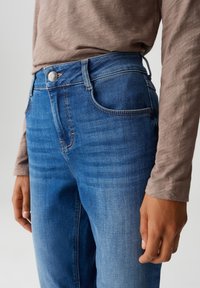 Persoon die blauwe spijkerbroeken en een lang, bruin bovenstuk draagt, weergegeven van de taille tot halverwege de dij met een ontspannen hand aan de zijkant.