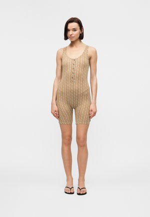 Γυναίκα στέκεται φορώντας ένα μπεζ ριγέ ολόσωμο σορτς χωρίς μανίκια με κουμπιά στο μπροστινό μέρος και μαύρα σαγιονάρια, σε λευκό φόντο.