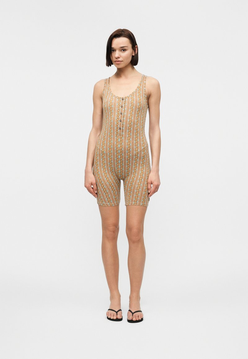 Γυναίκα στέκεται φορώντας ένα μπεζ ριγέ ολόσωμο σορτς χωρίς μανίκια με κουμπιά στο μπροστινό μέρος και μαύρα σαγιονάρια, σε λευκό φόντο.