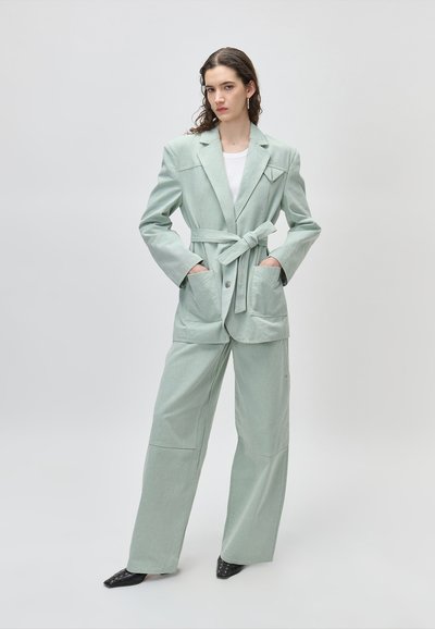 Γυναίκα όρθια με τα χέρια στις τσέπες, φορώντας ανοιχτά πράσινο σακάκι με ζώνη και αντίστοιχο παντελόνι με φαρδιά πόδια, μαύρα μυτερά παπούτσια και λευκό τοπ.