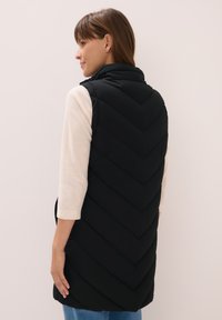 Schwarze Pufferweste mit einem Chevron-Steppdesign. Hoher Kragen und seitliche Taschen. Das Modell trägt einen hellen Pullover und Denim-Underteile.