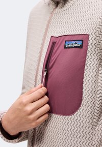 Χέρι κλείνει το μωβ τσέπη στο στήθος σε μπεζ fleece jacket Patagonia με ζιγκ-ζαγκ σχέδιο και εμπρόσθιο φερμουάρ.