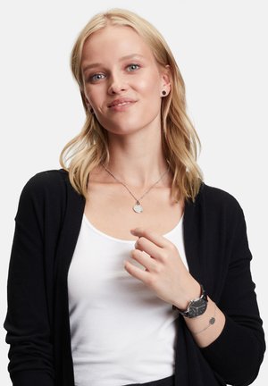 Silberne Pendants-Neklace an einer Frau, die einen schwarzen Cardigan über einem weißen Oberteil trägt. Schwarze Uhr an ihrem Handgelenk mit silbernen Akzenten.