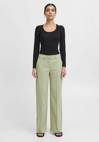Schwarzer langärmeliger Body, hellgrüne hoch taillierte weit geschnittene Hose mit vorderen Falten und schwarze Sandalen mit offenen Zehen. Schlichte runde Ohrringe.